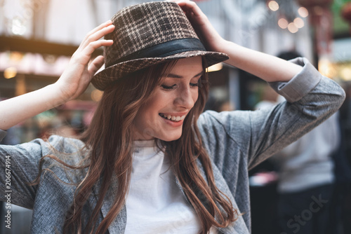 Attractive young woman is laughing outside. She is touching with hands trendy hat on head and looking down with joy