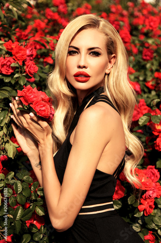 sexy woman with blond hair in luxurious dress posing in spring garden with flowering rose bushes