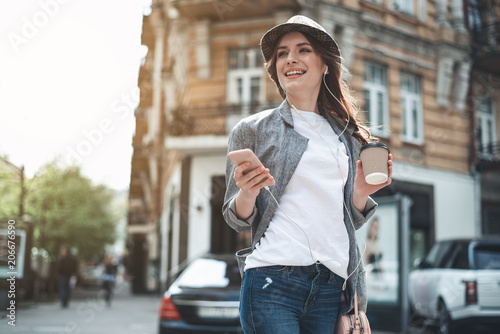 Cheerful young woman in trendy hat and earphones is standing in the street. She is holding smartphone and hot drink while looking away