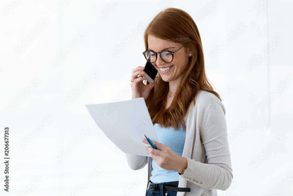 Beautiful young business woman using her mobile phone and reviewing papers in the office. Beautiful young business woman using her mobile phone and reviewing papers in the office.