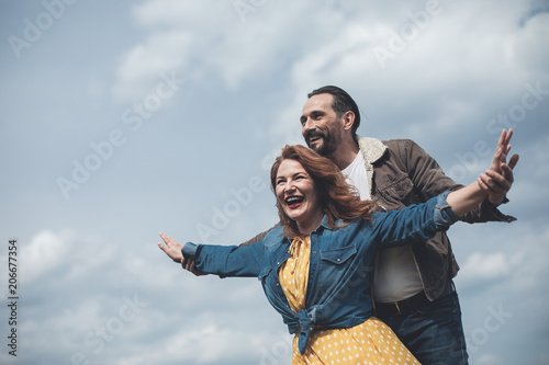 I am free. Portrait of excited woman is stretching arms sideways while man is embracing her from behind. They are enjoying wind and sky together 