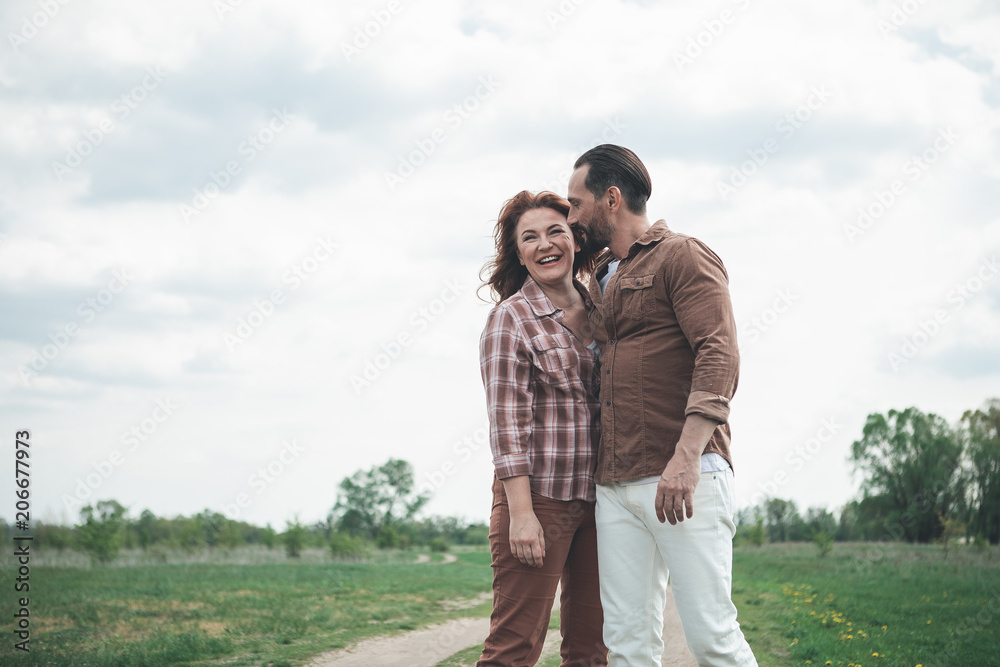 Portrait of glad middle-aged woman enjoying hug of her husband. She is smiling while lovers are standing on green field Portrait of glad middle-aged woman enjoying hug of her husband. She is smiling while lovers are standing on green field