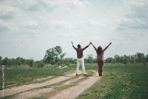 We are free. Joyful loving couple is standing on grassland path with relaxation. They are holding and stretching hands up with excitement. Focus on their back 
