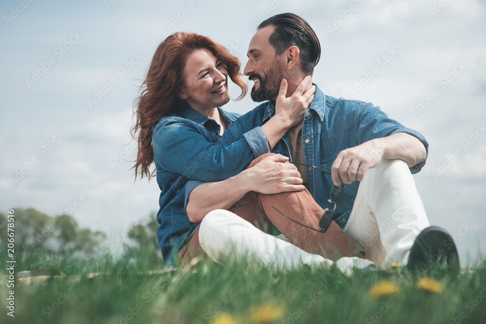 Low angle of happy middle-aged husband and wife are sitting on grass with relaxation. They are cuddling and smiling Low angle of happy middle-aged husband and wife are sitting on grass with relaxation. They are cuddling and smiling