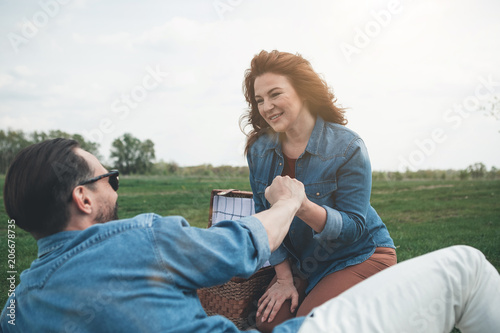Cheerful loving couple is holding hands with gentleness while relaxing on the meadow. Lady is looking at husband with love and smiling 