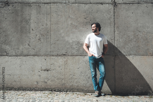 Happy alone. Full length portrait of young bearded male person standing against concrete wall. He is looking aside with smile. Copy space in left side