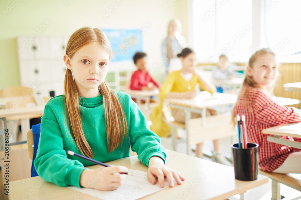 Serious and clever elementary schoolgirl looking at you while carrying out written task at lesson