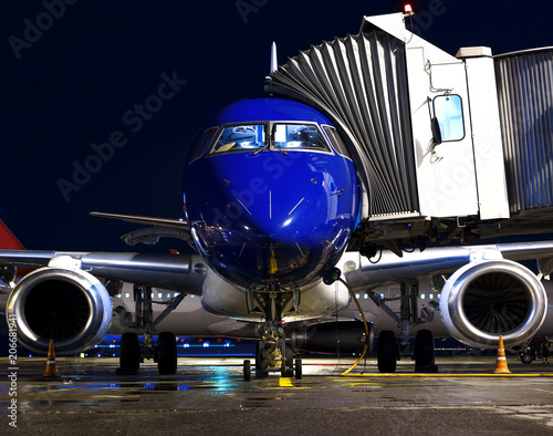 blue plane with a passenger tunnel parked at the gate at night in the background of another plane in the back