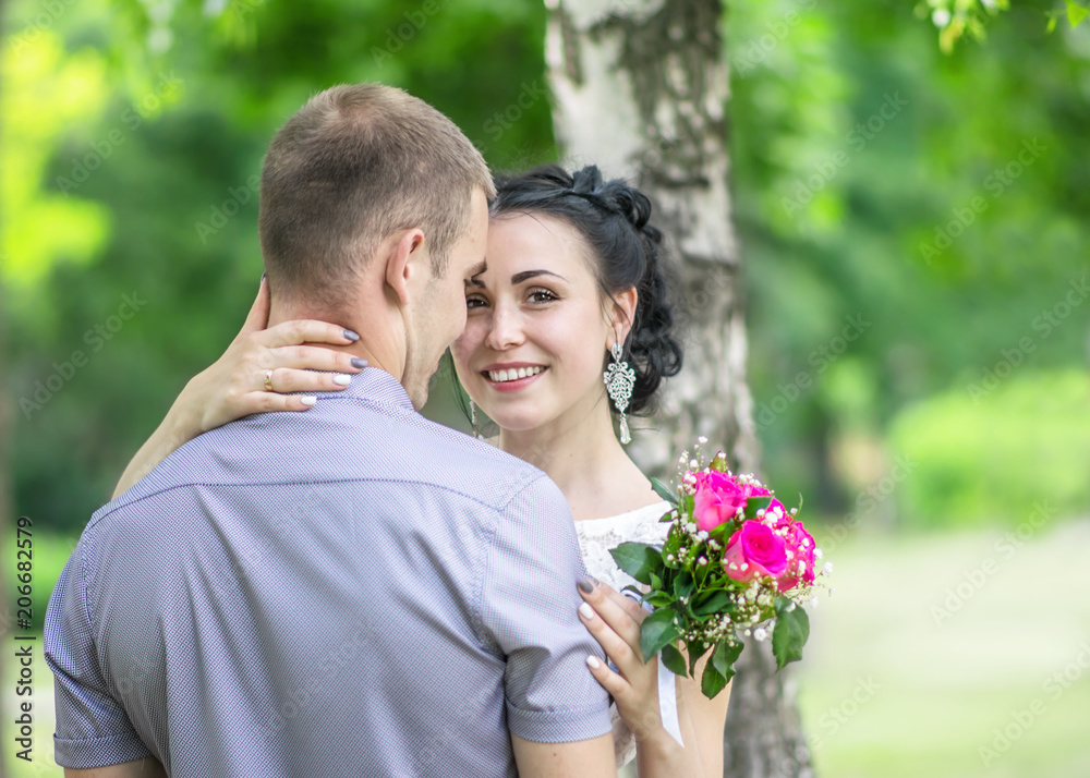 Portrait of a beautiful young female bride with small wedding pink flower roses bouquet smiling, gently hugging bridegroom neck and looking into camera at sunny summer park.