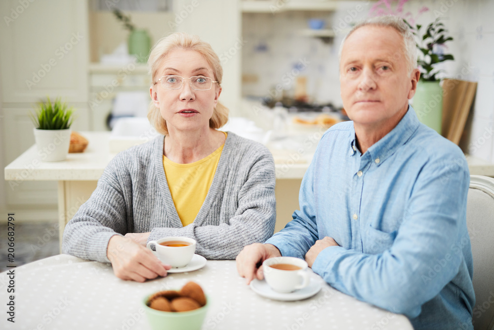 Retired couple looking at you while sitting by table and having tea with cookies in the kitchen Retired couple looking at you while sitting by table and having tea with cookies in the kitchen