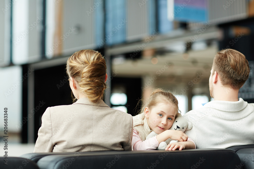 Obraz premium Cute little traveler looking at camera while sitting between her parents in airport lounge