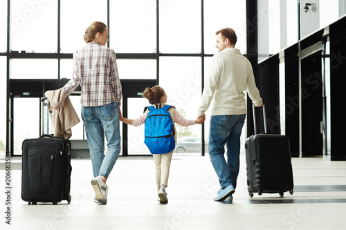 Back view of parents and little girl with baggage walking along airport lounge after arrival