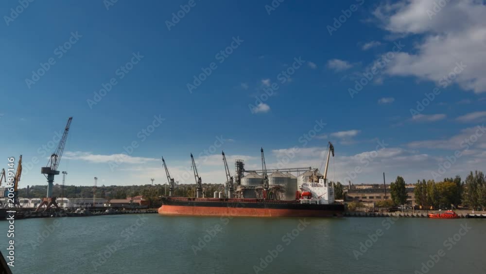 Timelapse of grain terminal at seaport on sunny day. Cereals bulk transshipment to vessel loading grain crops on bulk ship from large elevators at the berth. Transportation of agricultural products. Timelapse of grain terminal at seaport on sunny day. Cereals bulk transshipment to vessel loading grain crops on bulk ship from large elevators at the berth. Transportation of agricultural products.