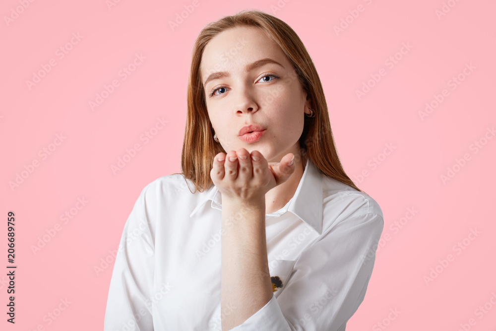 Attractive female with blue eyes, healthy skin, blows air kiss, expresses her love to close people, dressed in elegant white blouse, isolated over pink background. Horizontal shot of lovely woman Attractive female with blue eyes, healthy skin, blows air kiss, expresses her love to close people, dressed in elegant white blouse, isolated over pink background. Horizontal shot of lovely woman