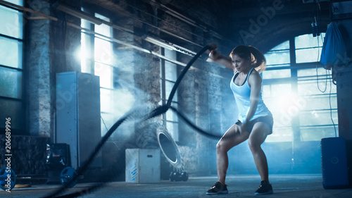Athletic Female in a Gym Exercises with Battle Ropes During Her Cross Fitness Workout  High-Intensity Interval Training. She s Muscular and Sweaty  Gym is in Deserted Factory. Cold Ambient.