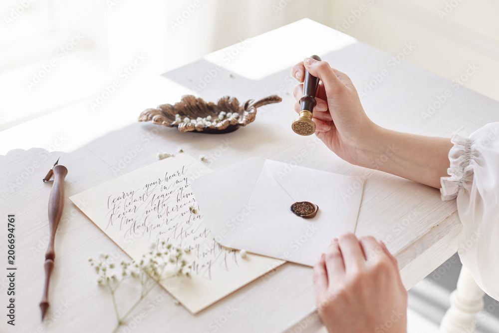 Obraz premium Girl writes a letter to her beloved man sitting at home at the table in a white light dress, purity and innocence. Curly blonde romantic look, beautiful eyes. White wildflowers on the table