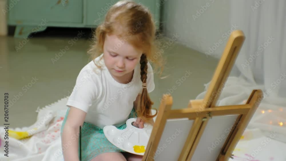 Beautiful red-haired girl sits on the floor in the studio in front of the easel and draws a brush and colored paints Beautiful red-haired girl sits on the floor in the studio in front of the easel and draws a brush and colored paints