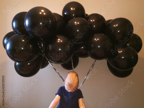 a small red-haired boy is delighted with the gift in the form of a huge cloud of black helium balloons presented for his birthday photo