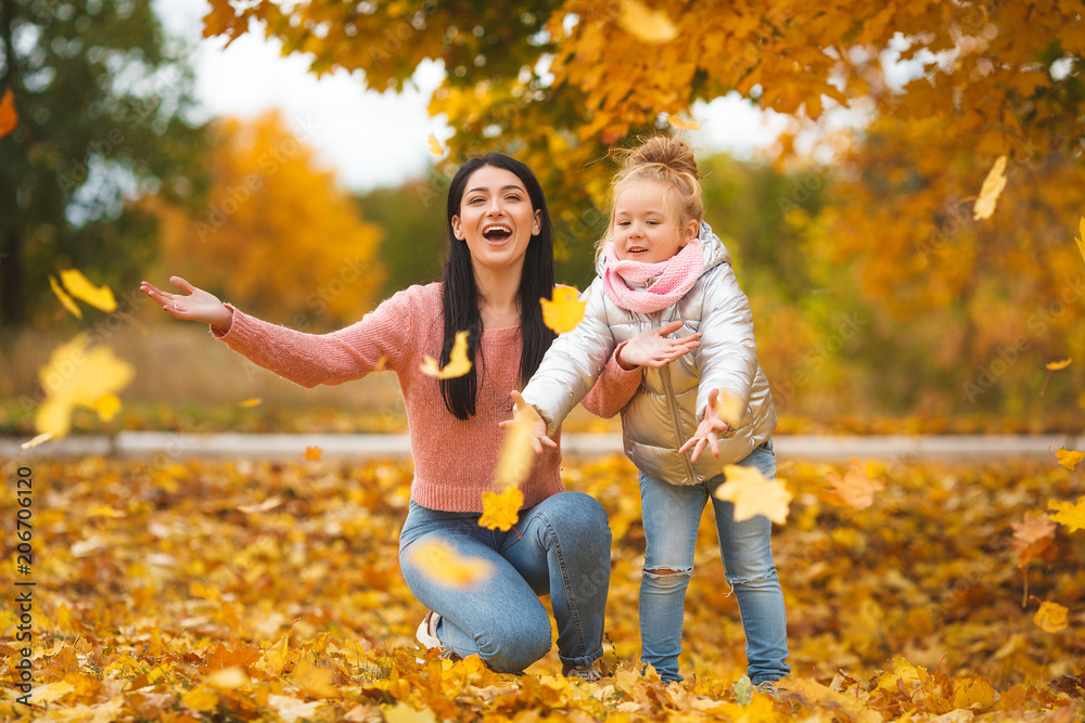 Fototapeta premium Cheerful girls playing with yellow leaves. Happy mother and little child in the fall time having fun