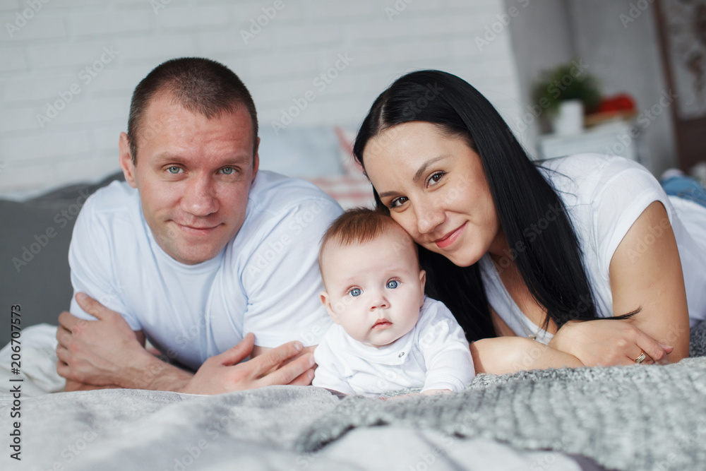 Fototapeta premium Young mom and dad and their baby son lie in bed and look at the camera indoors. The concept of a happy family