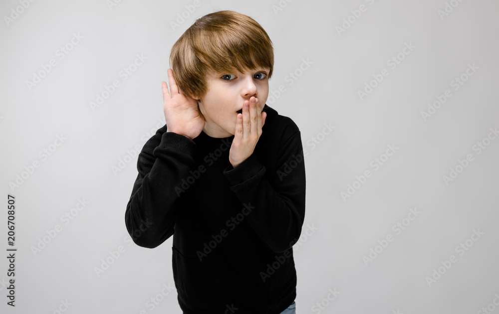 Charming teenager with blond hair and dark eyes. The boy closes his mouth with one hand, brings his other hand to his ear Charming teenager with blond hair and dark eyes. The boy closes his mouth with one hand, brings his other hand to his ear