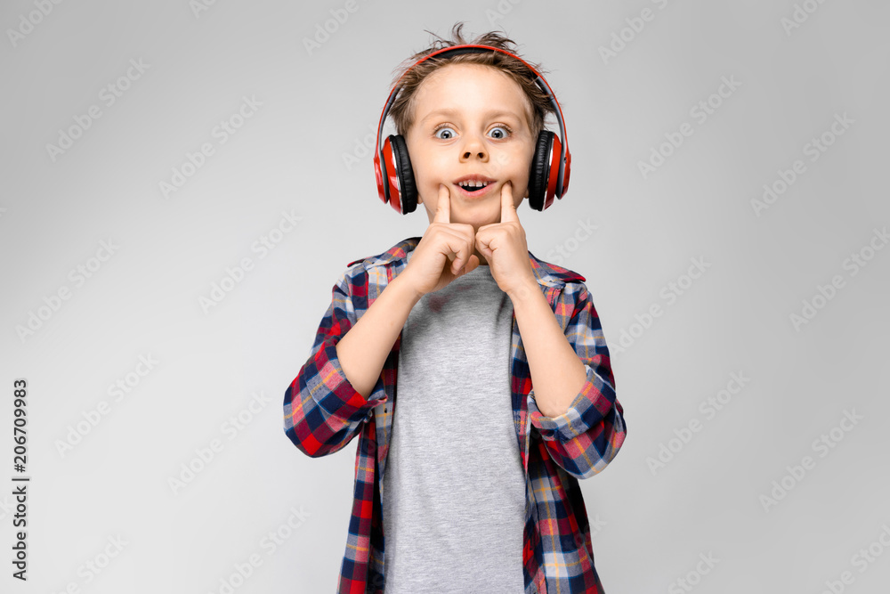 A handsome boy in a plaid shirt, gray shirt and jeans stands on a gray background. A boy in red headphones. The boy stretches his fingers with a smile. A handsome boy in a plaid shirt, gray shirt and jeans stands on a gray background. A boy in red headphones. The boy stretches his fingers with a smile.