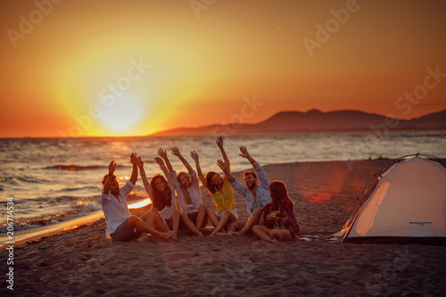 Happy friends sitting on the beach singing and playing guitar during the sunset