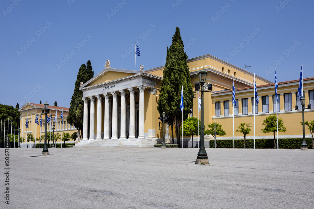 Greece, Athens: Front view of famous Zappeion building in the city center of the Greek capital and part of National Gardens with national flags and blue sky in the background - concept architecture.