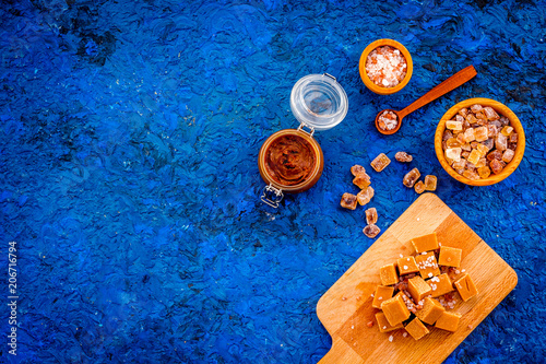 Salted caramel background. Melted caramel in glass jar, caramel cubes on cutting board, sugar and salt on blue desk top view copy space