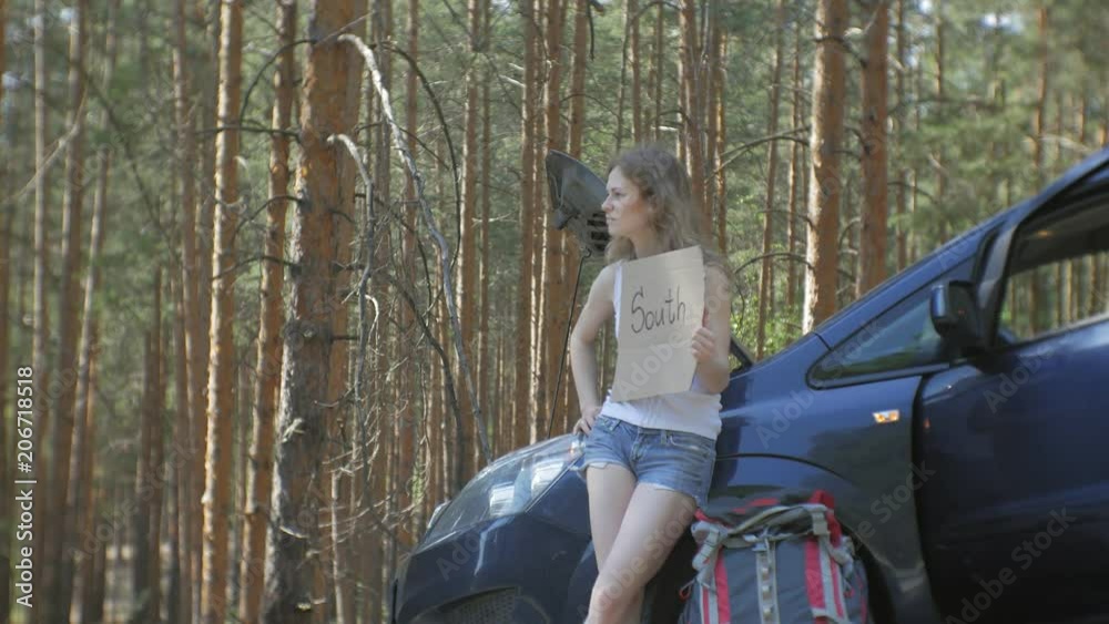Young beautiful woman hitchhiking standing on the road with a backpack on a table with an inscription SOUTH near a broken car Young beautiful woman hitchhiking standing on the road with a backpack on a table with an inscription SOUTH near a broken car