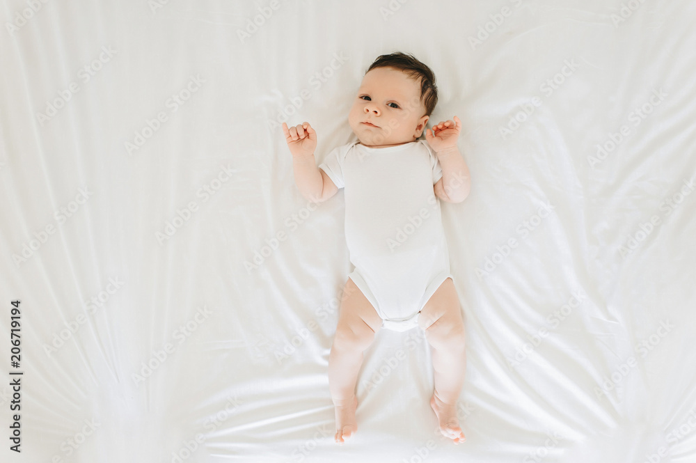 overhead view of cute newborn baby in white bodysuit lying on bed overhead view of cute newborn baby in white bodysuit lying on bed