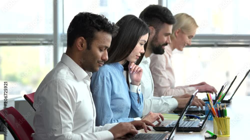 Smiling tech support workers typing on laptop. Four smiling clerks sitting beside each other and working on laptops. Smiling tech support workers typing on laptop. Four smiling clerks sitting beside each other and working on laptops.