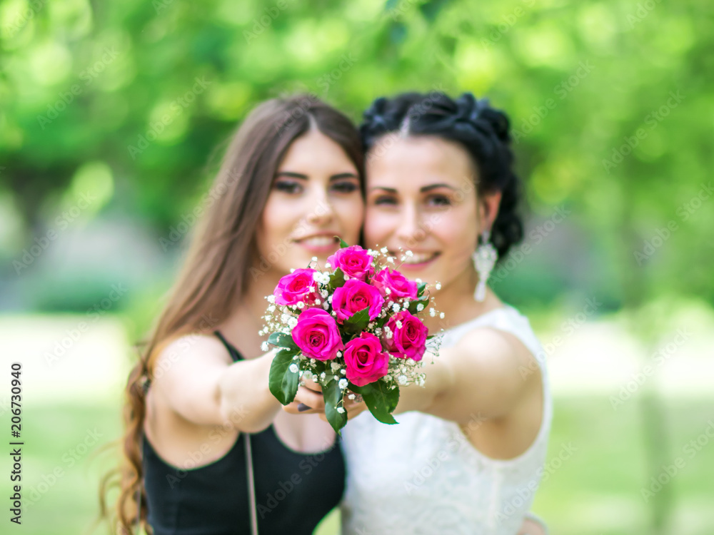Defocused portrait of two beautiful young women holding together wedding bouquet and looking into camera at green park. Bride and bridesmaid holding bouquet on outstretched arms. Focus on the flowers. Defocused portrait of two beautiful young women holding together wedding bouquet and looking into camera at green park. Bride and bridesmaid holding bouquet on outstretched arms. Focus on the flowers.