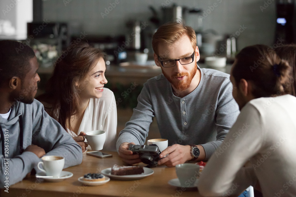 Young redhead guy holding camera showing photos sharing recent memories or travel impression hanging with diverse friends, smiling multiracial millennial people talking sitting together at cafe table