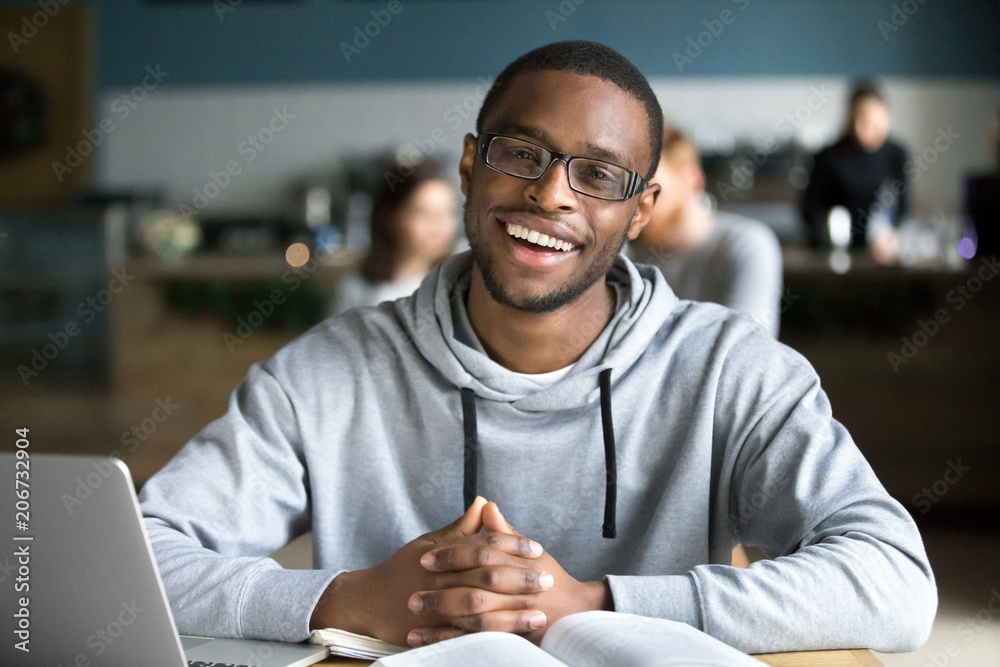 Happy african casual man in glasses sits at cafe table looking at camera, cheerful black student posing studying with book in coffee house, smiling businessman works on laptop in cafeteria, portrait Happy african casual man in glasses sits at cafe table looking at camera, cheerful black student posing studying with book in coffee house, smiling businessman works on laptop in cafeteria, portrait
