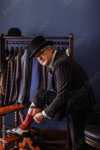 Welthy elderly male businessman in expensive suit resting one leg on antique stool in victorian style fixing his sock on at tailor's workshop. photo