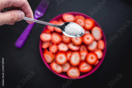 Fototapeta Naklejka Na Ścianę i Meble -  Top view of a teaspoon of sugar and sliced ​​fresh red strawberries and a plastic fork. Fruit arranged on a purple plate, ready to eat. Sprinkling strawberries with sugar.