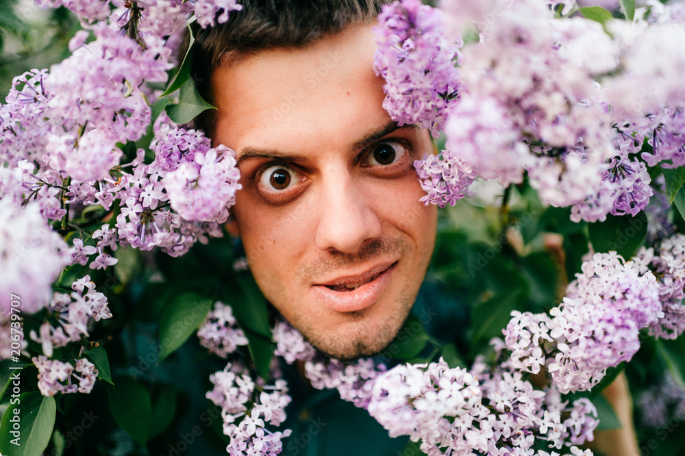 Obraz premium Closeup odd unusual male portrait outdoor at nature in beautiful lilac bushes. Adult funny man face grimace surrounded by blooming flowers. Emotions and facial expxression. Clowning and fooling.