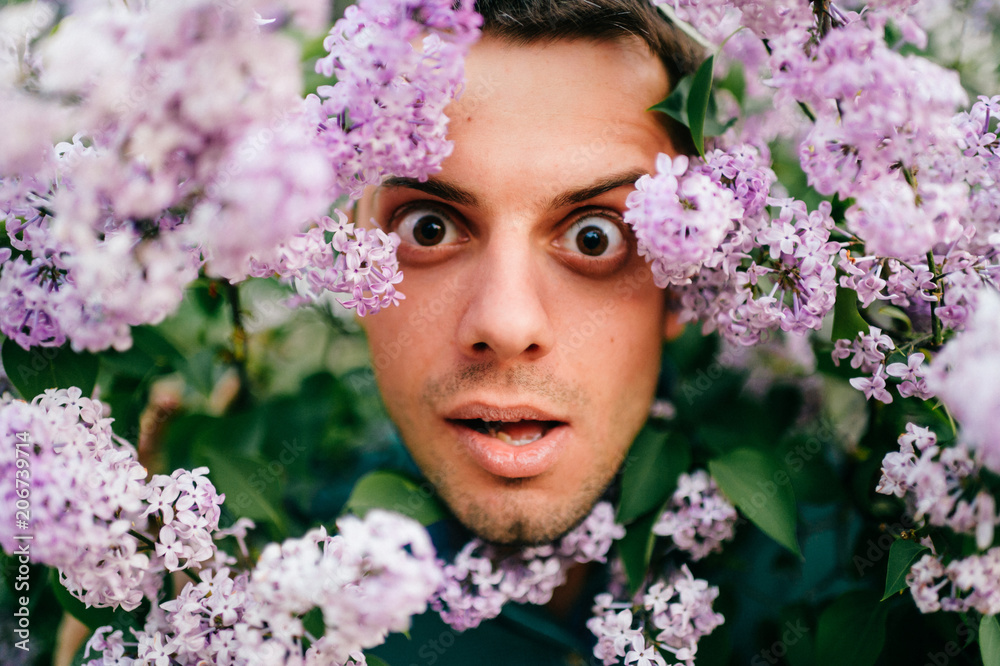 Obraz premium Closeup odd unusual male portrait outdoor at nature in beautiful lilac bushes. Adult funny man face grimace surrounded by blooming flowers. Emotions and facial expxression. Clowning and fooling.