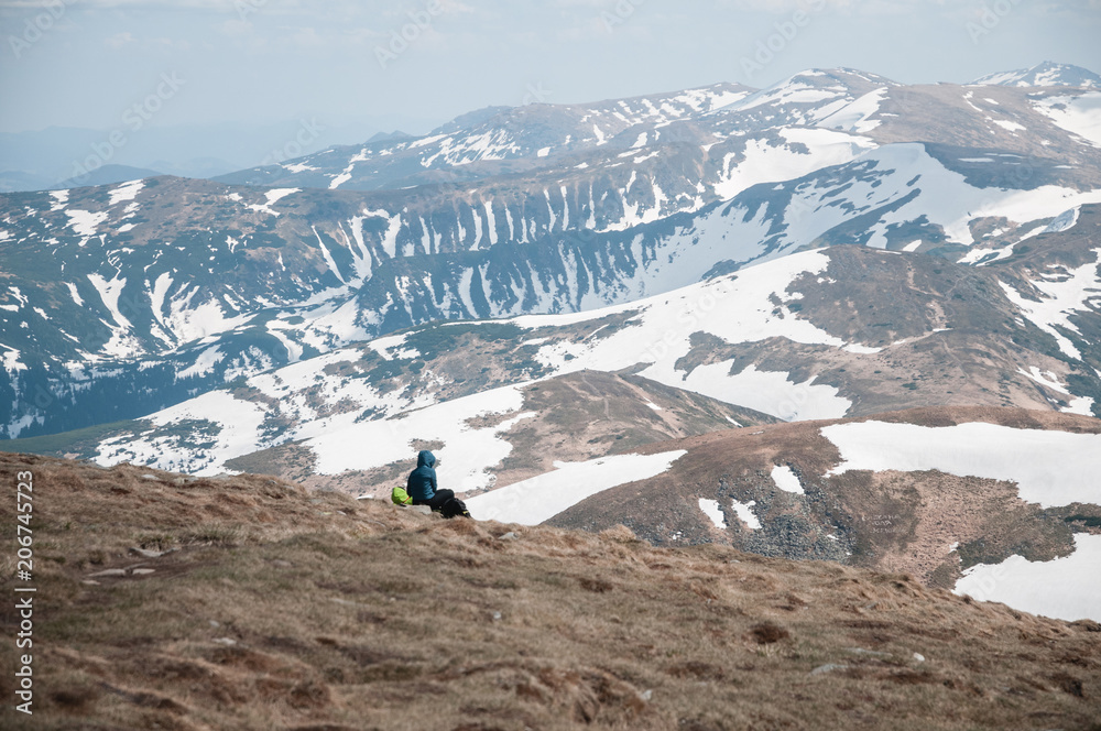 The man sits on top and looks at the snow-capped peaks of the mountains in the distance on a clear day in light clouds. Carpathians, Goverla, Ukraine.