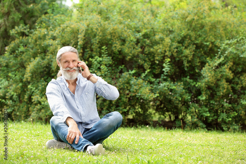 Handsome mature man with mobile phone sitting on green grass in park