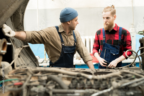 Young male car mechanic standing by broken truck and consulting with experienced bearded colleague about repair