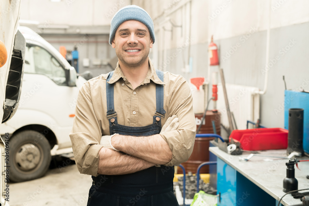 Portrait of young Caucasian male auto mechanic standing in repair shop with crossed hands and smiling at camera cheerfully