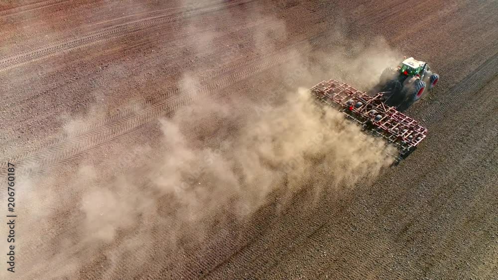 Big farm tractor tilling dusty Springtime fields, aerial view.
