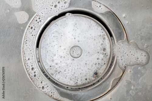 White foam with bubbles of cleane in a washbasin. Drain hole with soap bubbles in metal sink, macro view. Mechanically adjustable drain plug closeup. photo