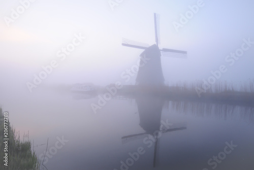 Windmill stands next to a canal and is reflected in the water in Netherlands in the fog of early morning.