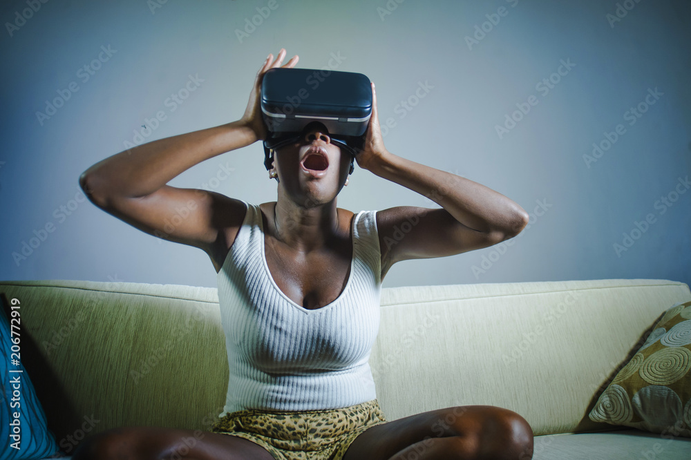young attractive black afro American woman playing amazed and surprised virtual reality video game wearing VR goggles at home living room couch