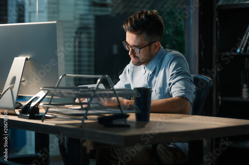 Obraz na plátně Young man in glasses working late alone at desktop in office taking notes and reading information