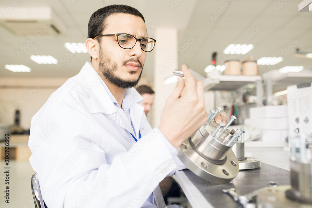 Serious frowning handsome young Arabian male engineer in lab coat viewing detail of bearing while assembling technical equipment in workshop Serious frowning handsome young Arabian male engineer in lab coat viewing detail of bearing while assembling technical equipment in workshop