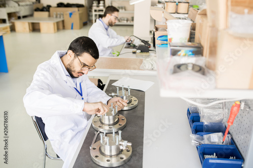 Concentrated busy young male engineer in lab coat and glasses focusing on bearing production and tightening bolts with hands while sitting at table